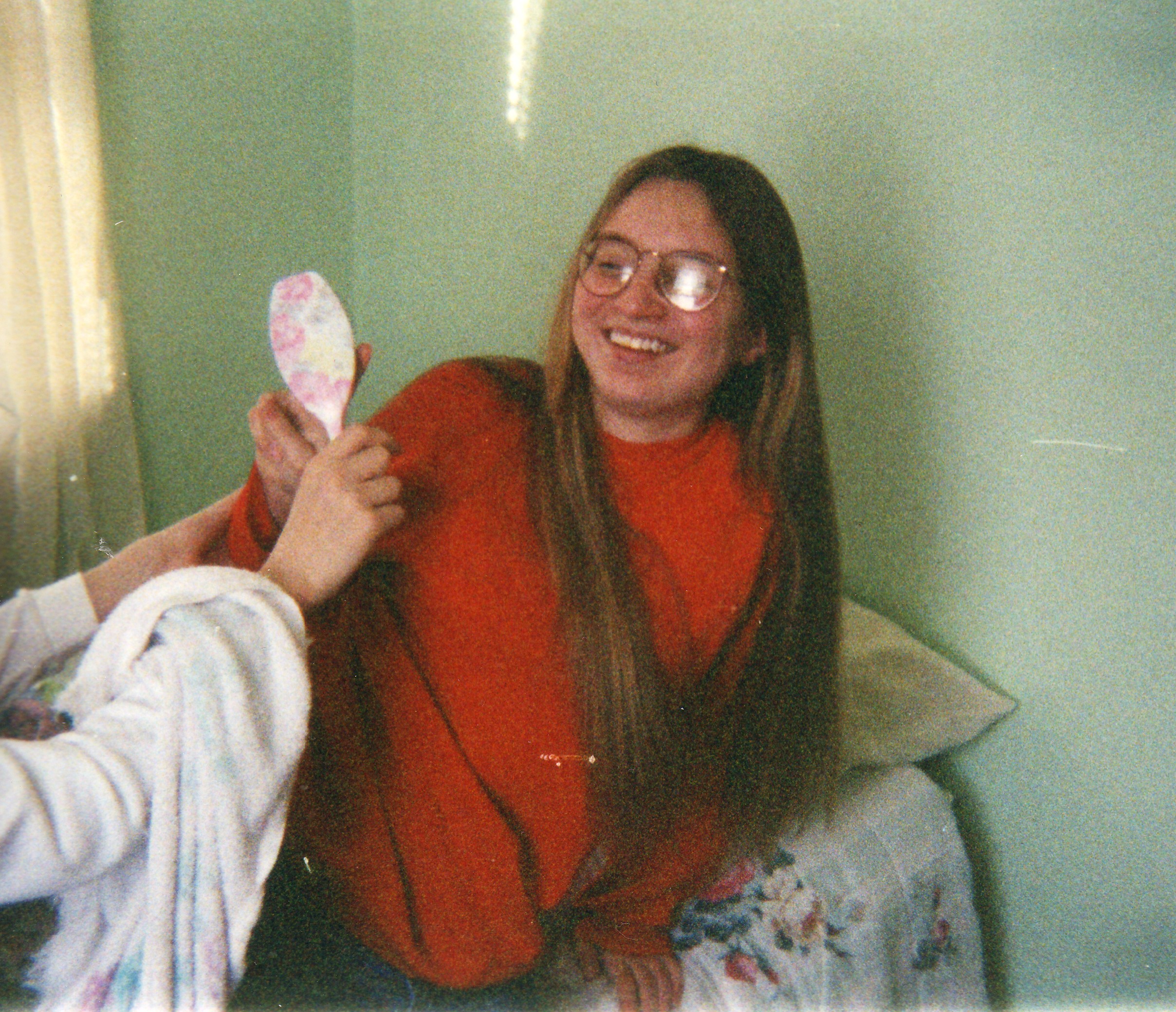 A teenage girl wearing a red sweater and wire rimmed glasses laughs while a hairbrush is waved in her face